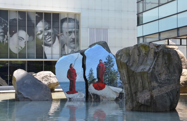 photograph or painting of a woman in red standing on a rock, directly onto the surface of a large rock in a shallow pool of water outside Ryerson Image Center, three large black and white photographs of people's heads are above and behind the artwork