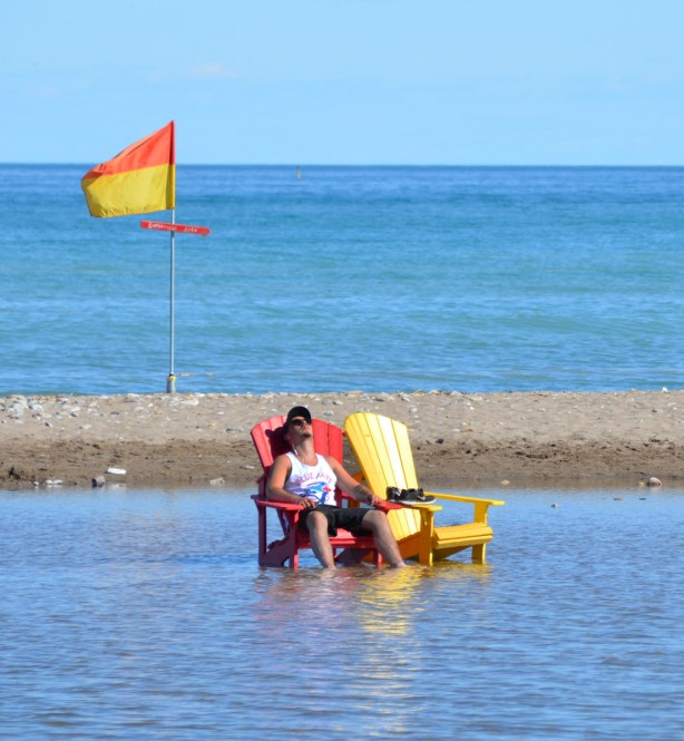 a man sits on a red Muskoka chair in the flooded part of Woodbine beach. A red and yellow flag flies behind him. 