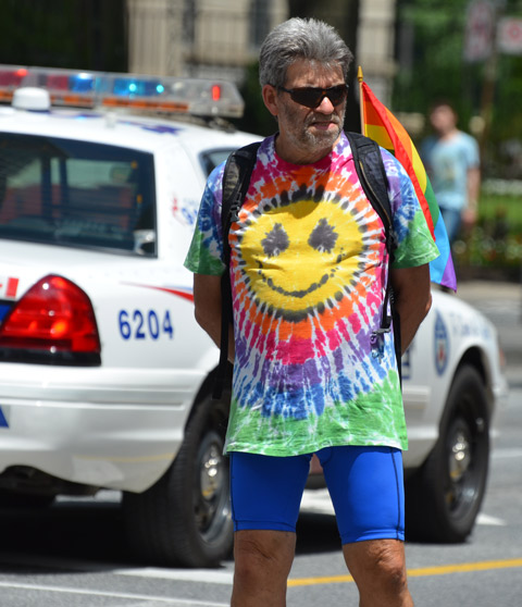 a middel aged man in a tie dyed shirt with a big happy face in the middle of it, stands by a police car as he watches the dyke march 