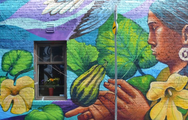 part of a larger mural on a wall with a window, a large painting of a woman appears to be looking at the window, a melon or gourd is in the mural too 