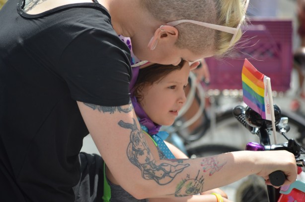 a mother leans over her young daughter who is sitting in front of her on a bike, Small rainbow flag is on the handlebars 