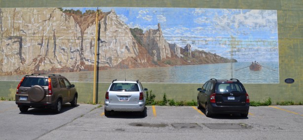 Three cars are parked in front of a large mural of the Scarborough Bluffs, there is a small row boat on Lake Ontario in front of the cliffs. 