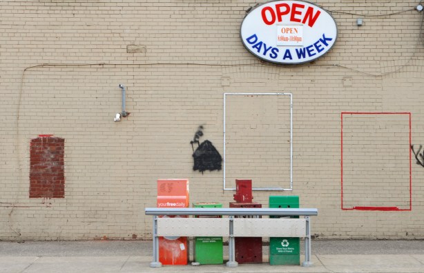 4 nespaper boxes lined up on a sidewalk in front of a beige wall, store, with sign that says open days a week. 