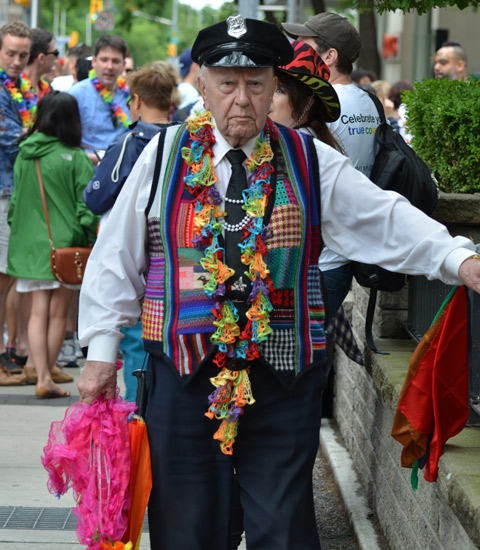 an older man is standing on the sidewalk, wearing a police cap and a patchwork vest in rainbow colours with all kinds of different fabrics, busy, bright, 