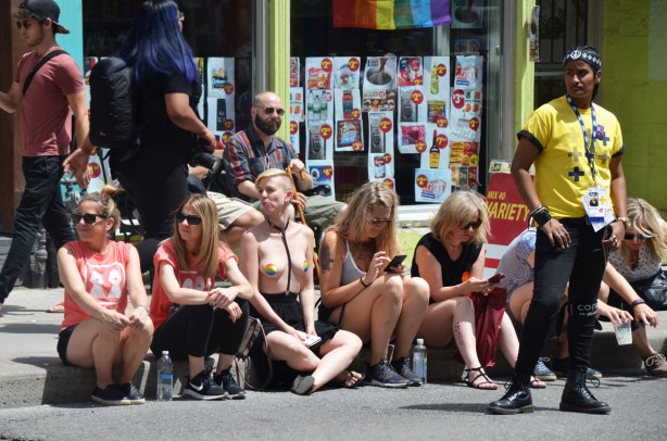 a group of people sittingo n the sidewalk on Yonge street as they watch the Dyke March. One woman is topless, two women are on cellphones
