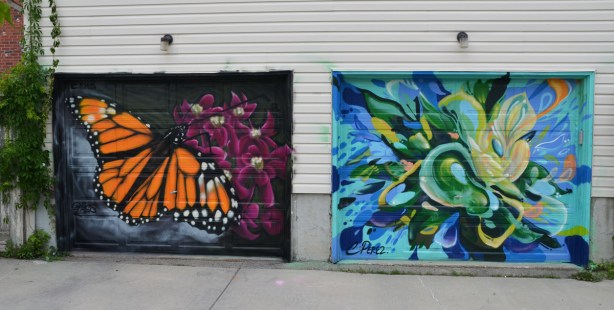 a mural on a garage door in an alley, part of butterflyways project - two adjacent garage doors, the left one is a monarch butterfly landing on pink flowers, on the right is a riot of blues and greens