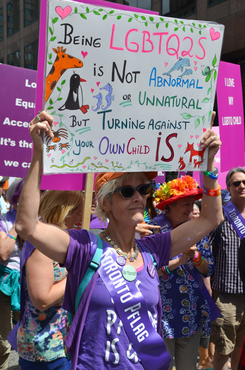 pflag member in purple, holding up a placard that say being LGBTQ2S is not abnormal or unnatural but turning against your own child is s 