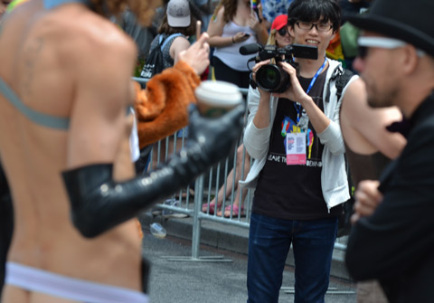 a young Asian man, photographer, smiles as he gets ready to take pictures of mostly nude man with long leather gloves on 