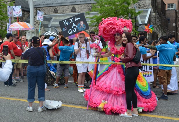 a man waiting for the pride parade to start holding a sign that says yup I am a gay. Person inlong frilly pink gown and massive pink feather head dress, posing for pictures