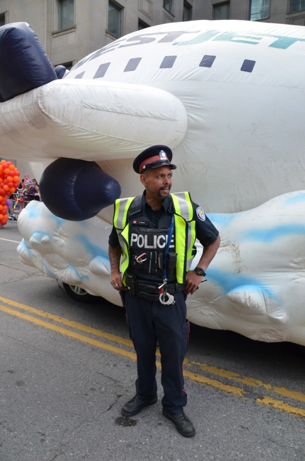 a police men watches an intersection of streets as the parade passes, he justs fits under the wing of a large inflatable westjet plane