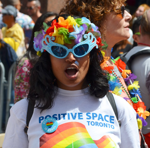 a woman wearing blue rimmed sunglasses and rainbow paper flowers in her hair is looking directly at the camera and shouting. She has a t shirt on that says Positive Space toroto 