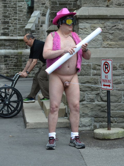 a man naked except for a pink fuzzy vest and a pink cowboy hat is standing on the sidewalk. There is a string tied around his penis. A man is pushing an empty wheelchair in the background