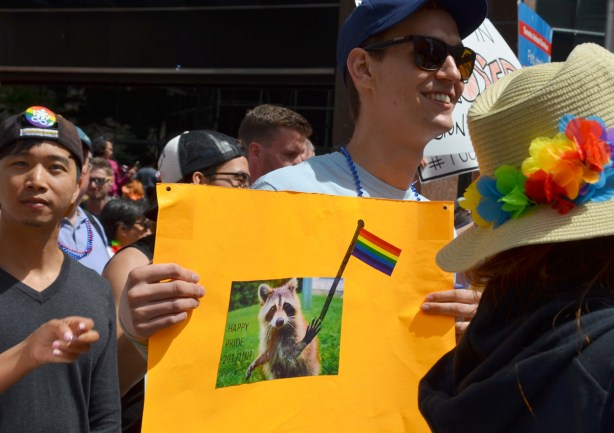 a young man holds an orange sign .. on the sign is a picture of a raccoon holding a rainbow flag with the words happy pride 2017