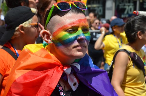 person with rainbow face paint, rainbow rimmed sunglasses and a rainbow flag draped over her back 