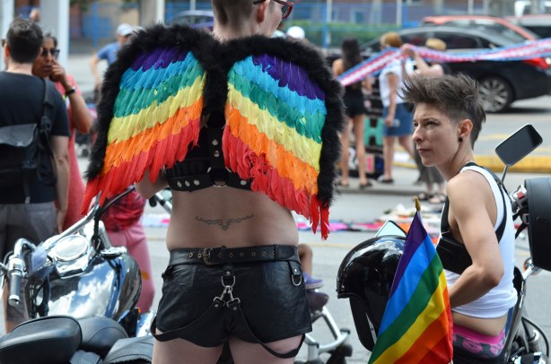 waiting for the dyke march to begin, two people by their motorcycle, one is in leather shorts and has angel wings made of rainbow coloured feathers