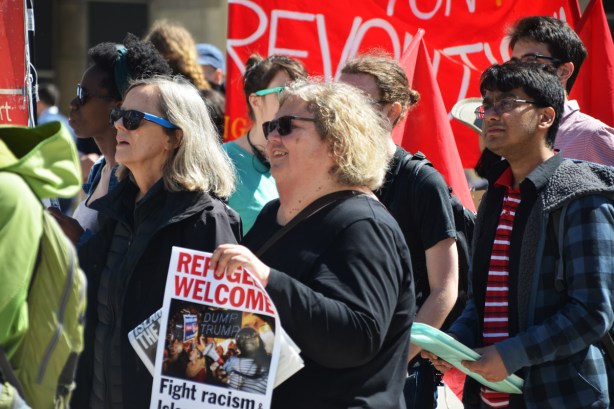 people walking in a protest, a large red and white banner in the background, a woman holding a sign that says refugees welcome with a photo of refugees on it