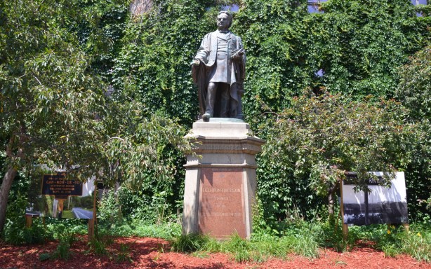 statue of Egerton Ryerson in a small garden with shrubs and small trees. Two large photographs also in the picture, one on each side of the statue