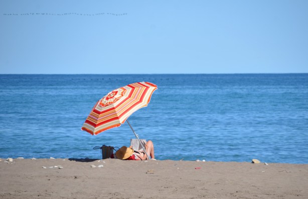 at the beach, a woman with a large yellow hat and red top lies under a red, orange and yellow striped umbrella, lake and sky in the background