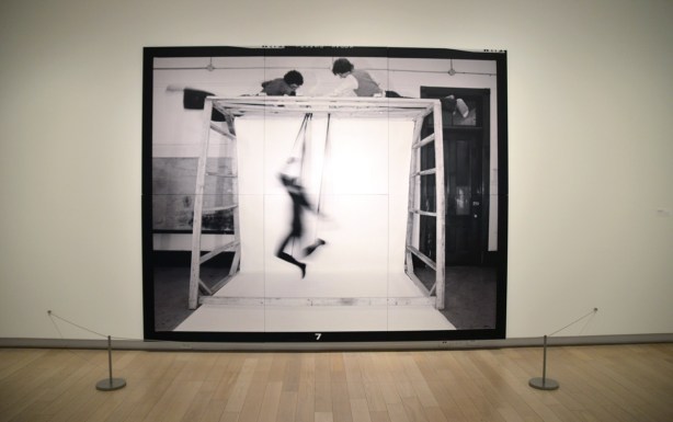 large black and white photograph in a gallery, two men on top of a large frame are controlling the movements of a human puppet