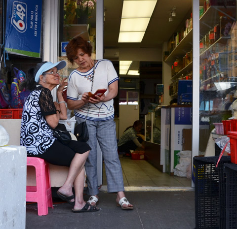 two women outside a store,looking at a phone, a woman inside is crouched on the floor, working. 