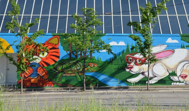3 small trees in front of a colourful mural by Uber5000 of animals, a green turtle by a blue creek, a white rabbit with red sunglasses and an orange tigerstriped kitten on the side of the THS building