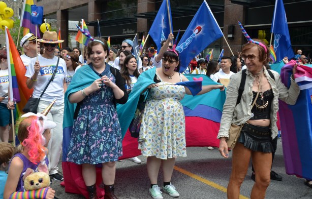 three women walking in the pride parade with a turquoise, blue and pink banner, and unicorn horns on their head, stop to talk to a young girl who also has a unicorn horn on her head