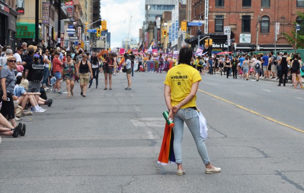 a volunteer wearing a yellow tshirt stands in the middle of yonge street facing the dyke march parade that has stopped just up the street, people are lining the sidewalks to watch the march 