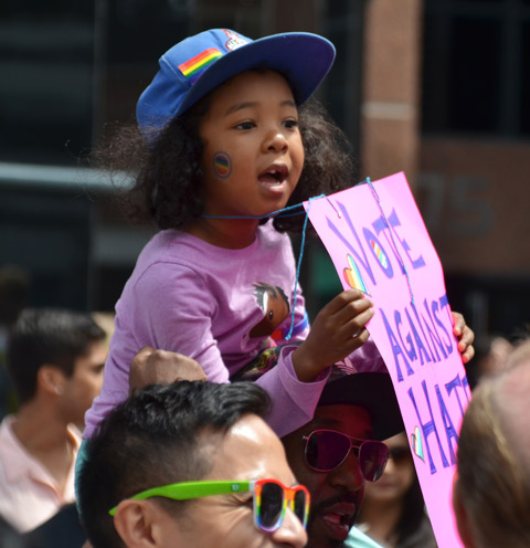 a young black girl is sitting on her father's shoulders. She is holding a pink sign with purple hand written letters that say vote against hate 