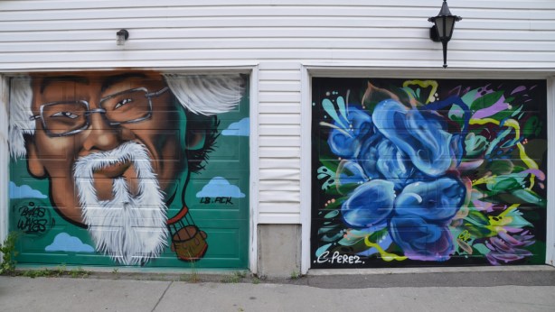 a mural on a garage door in an alley, part of butterflyways project - two adjacent garage doors, the left one is painted by braes and is a portrait of David Suzuki. On the right is a blue mural by C. Perez