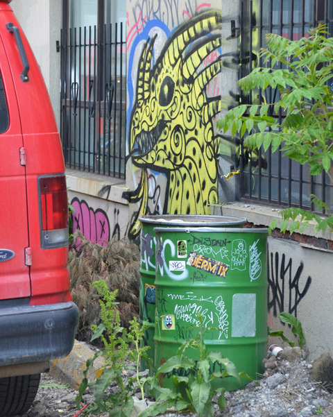 behind a red van and two green metal barrels, a yellow monster street art painting on a wall 