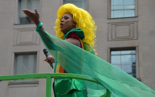 up on the TD float in the pride parade, a person is a long wavy yellow wig and wearing a striped top and short green shiny skirt, is blowing kisses to the crowd.