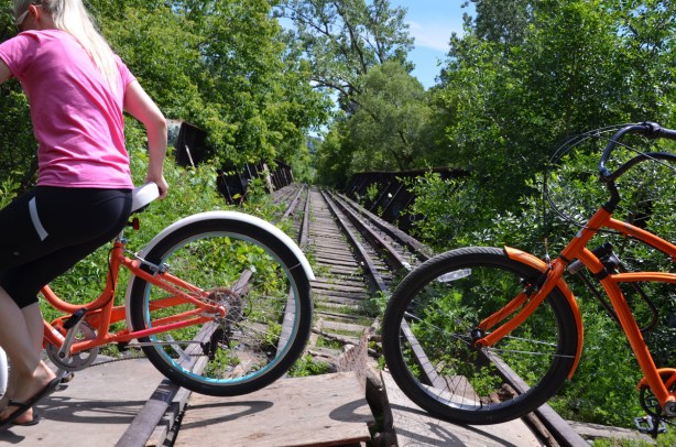 two cyclists walk their bikes across loose pieces of plywood over unused railway tracks 