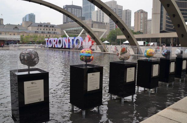 a line of sculptures on display, podius standing in the water of the fountain, arches, and 3D Toronto sign in the backgruond. 