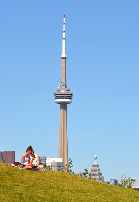 couple, man and woman, sitting together, on a grassy hill. The CN Tower is behind them. 