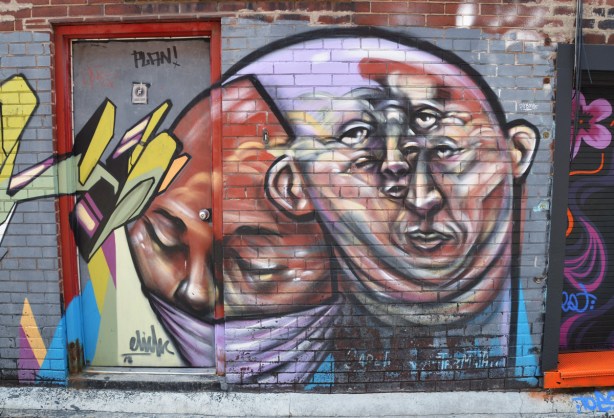 elicser street art piece in a lane, a face with another face with multiple eyes that might be part of the first's head or might be separate 