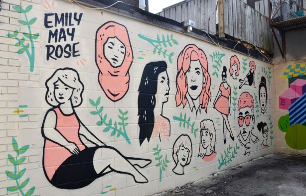 mural by emily may rose of various women's faces, in black and white and pink, long black hair, pink head scarf, pink dress, short hair, 