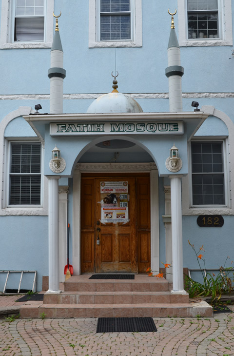 front of Fatih mosque, a light blue building with a small porch, two short minarets with gold cresents on top, a brown door, 
