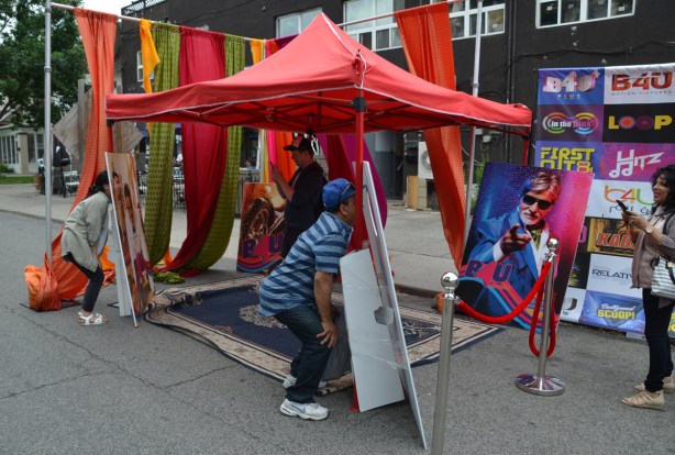at a street festival, two people are putting their heads through cut outs while others take their picture, cut outs are on faces of Bollywood actors and actresses.