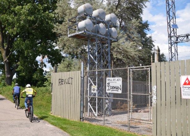 two boys ride bikes past the Mill Street Junction hydro station, fenced in area with danger signs, 