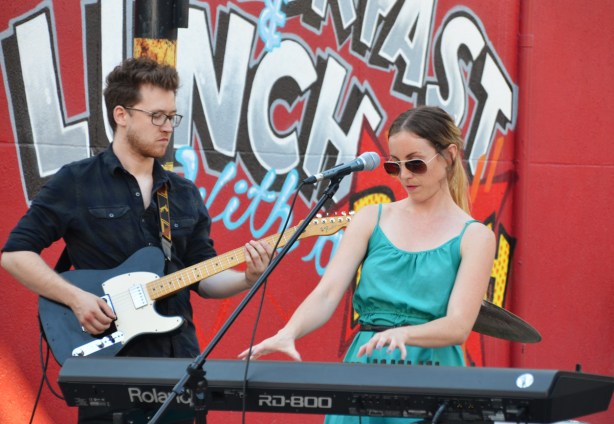 a woman in sunglasses and teal dress plays the keyboards. A man with a small beard is playing the guitar beside her, a red wall with text street art is behind them. 