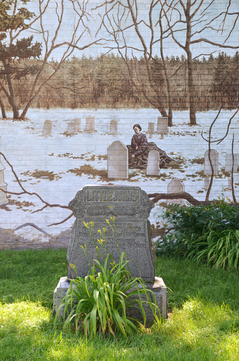 a real tombstone, surname Littlejohns, in a cemetery, with a mural in the background showing a woman kneeling by a grave in the winter, small amount of snow, no leaves on the trees 