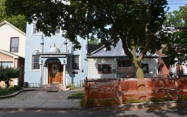 looking across a street, sidewalk, large tree with orange plastic fence around it to protect it during construction, a pale blue building that is a mosque and a small single storey house that is about to be demolished. 