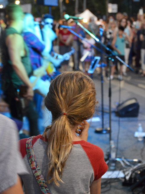 back of a girls head, long hair tied in a ponytail, outside, in the evening, as she watches a music performance with a small crowd of people 