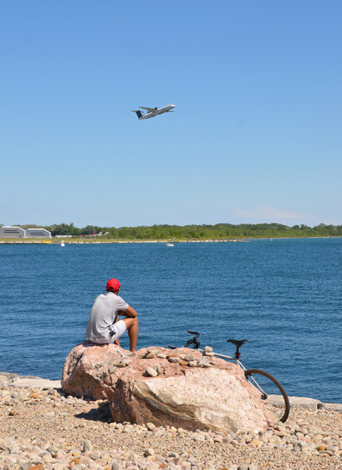 a man in a red baseball cap sits on a rock, his bike parked beside, while watching a pOrter airlines plane take off from Billy Bishop Airport 