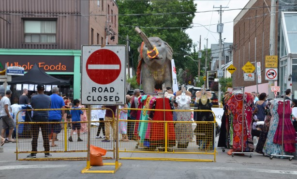 road losed sign and yellow metal barricades, street festival going on behind the sign & barricade. Also racks of sarees for sale and in the distance, a large fake elephant