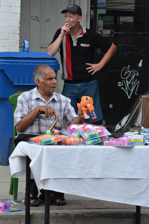 an older man sits behind a small table covered with a white cloth. on the table are toys that he is selling. he is holding an orange Nemo shaped bubble maker plastic toy. behind him, a man is standing smoking a cigarette