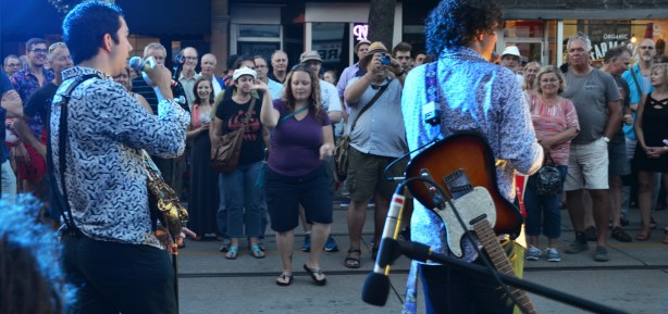 two musicians with lights on them, from the back, with the crowd standing on the street in front of them, one woman dancing 