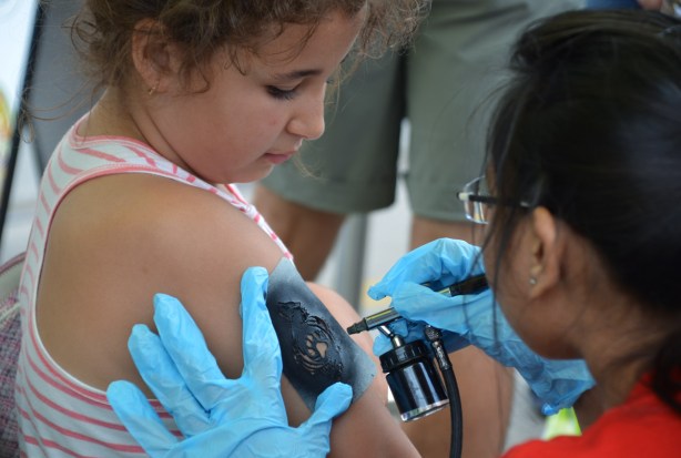 a woman is airbrushing black paint onto a stencil on the upper part of a small girls arm, temporary tattoo