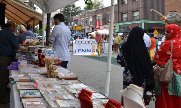 street scene at South Asian festival. a man hits a ball with a cricket bat, two women in head scarves are talking, men behind a table are selling jewellery and clothes 