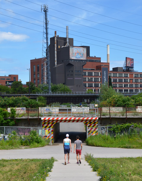 two men walk through a park towards an underpass under a railway track, factory in the background. 